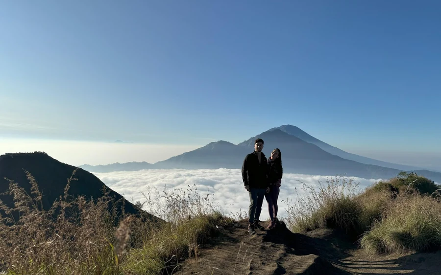 A couple standing on a ridge above a sea of cloud with a distant cone behind them