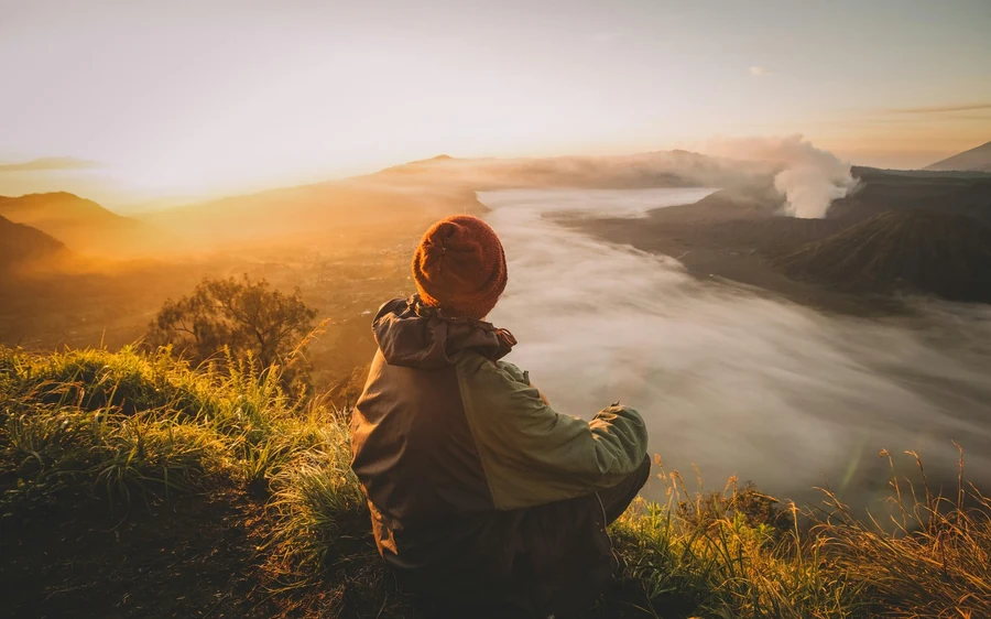 A person in a woolly hat sitting on the grass above a valley filled with morning mist