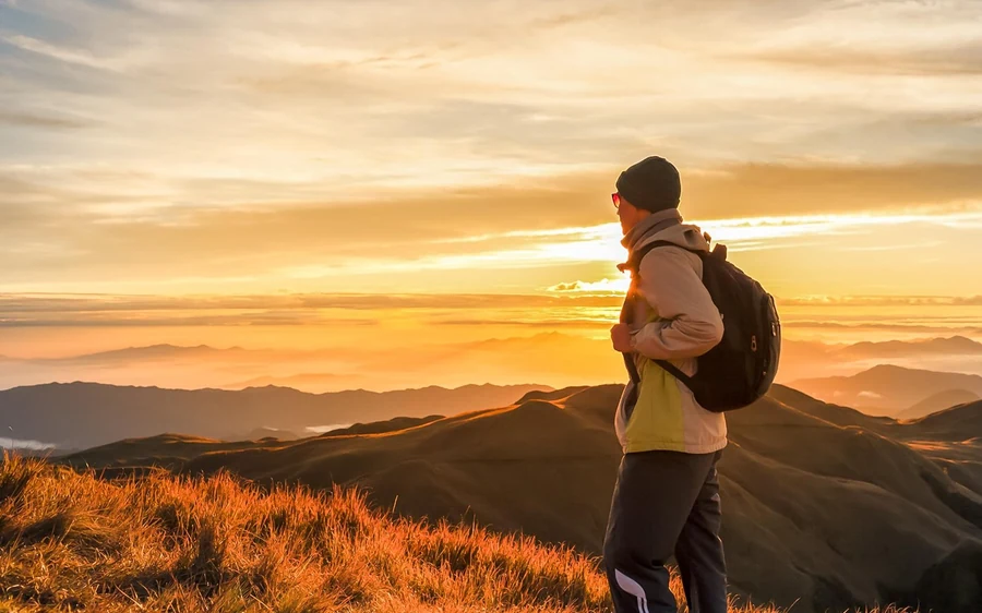 A hiker in a beanie with a backpack watching a gold sunrise over layered ridges