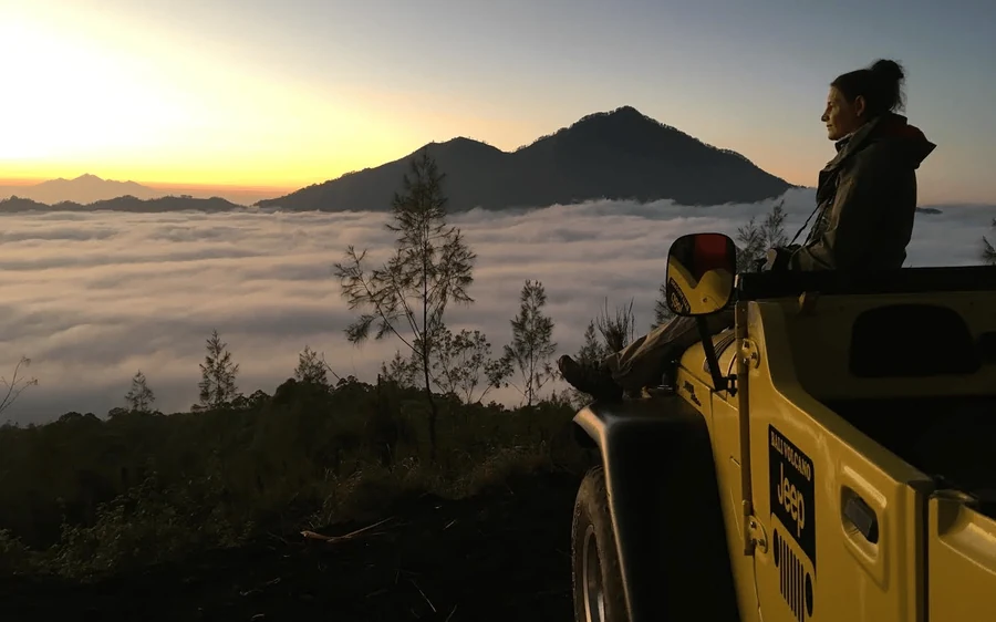 A woman sitting on a yellow jeep above a sea of cloud at first light