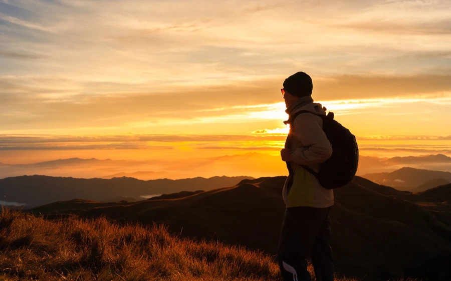 A hiker in a beanie watching a gold sunrise from a grassy ridge