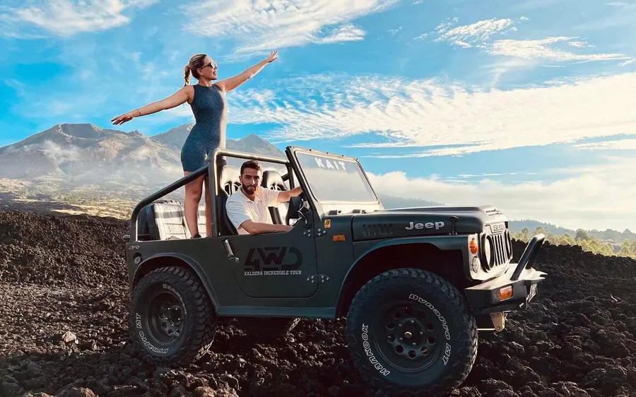 A woman standing up in a dark green jeep on black lava rock with the mountain behind