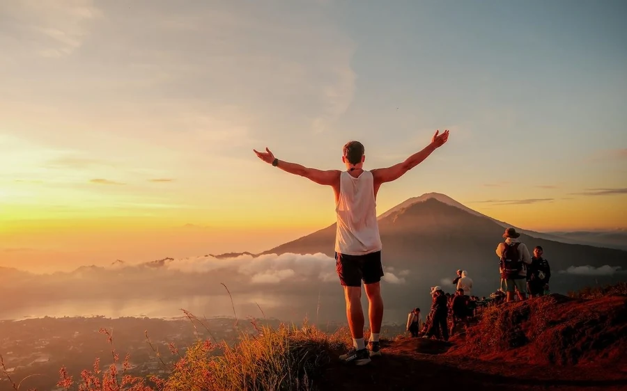 A man standing with his arms out on a summit at sunrise with a cone and a lake behind him