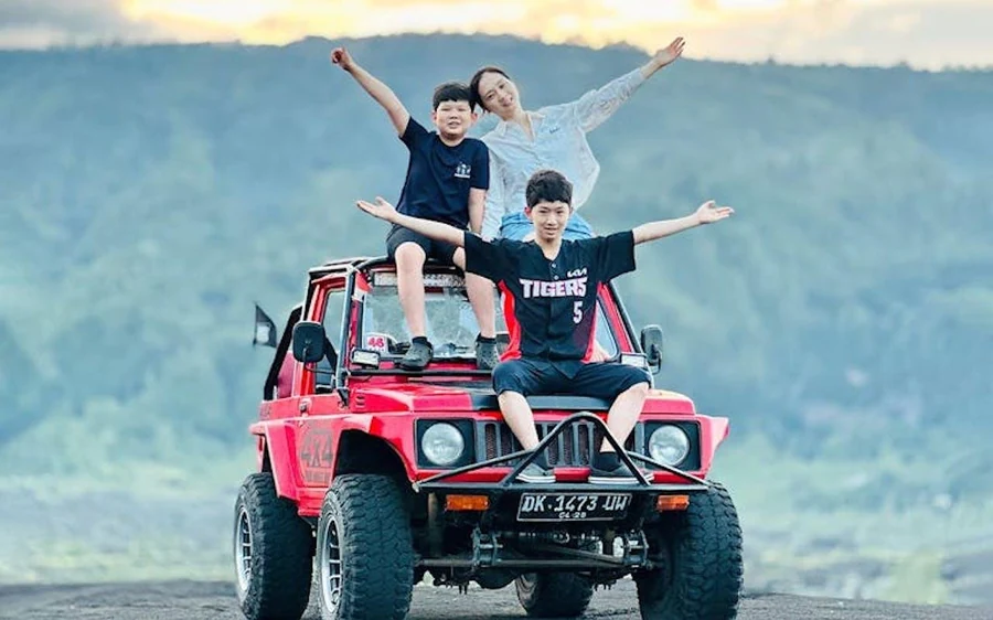 A family sitting on a red four-wheel drive with green crater slopes behind them
