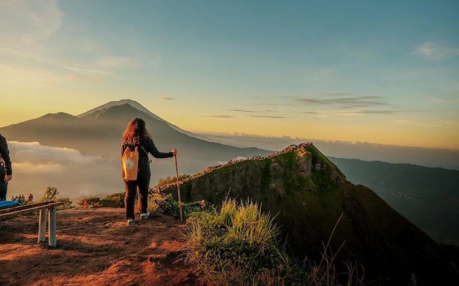 A woman with a walking stick and a yellow backpack looking towards a cone at dawn