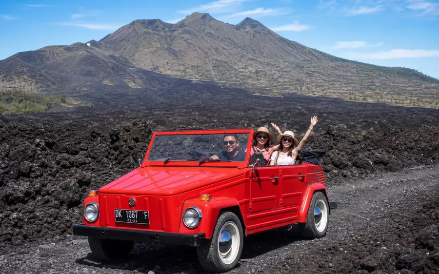 Three people waving from an open red classic VW on black lava with the mountain behind